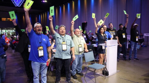 A row of people stand and raise bright green FOR cards beside a clear acrylic floor microphone, a lit number 7 microphone marker glowing overhead; visible credentials include DELEGATE and NATIONAL OFFICER.