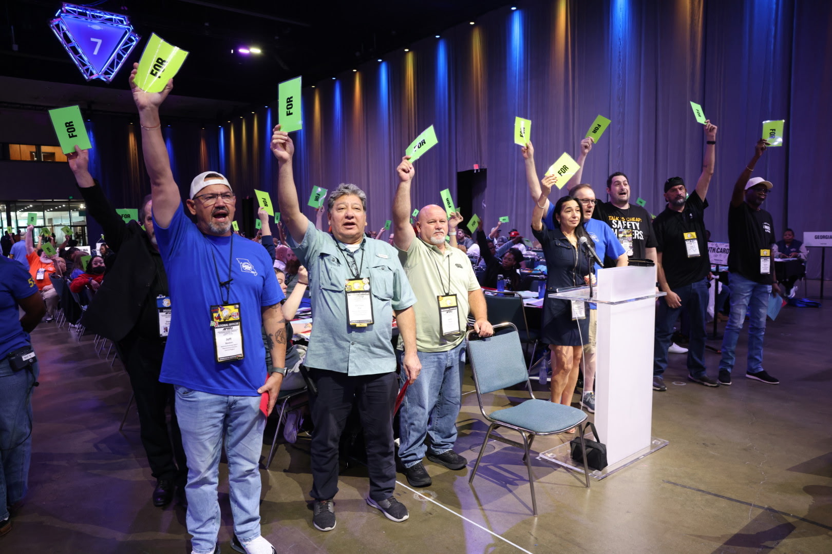 A row of people stand and raise bright green FOR cards beside a clear acrylic floor microphone, a lit number 7 microphone marker glowing overhead; visible credentials include DELEGATE and NATIONAL OFFICER.